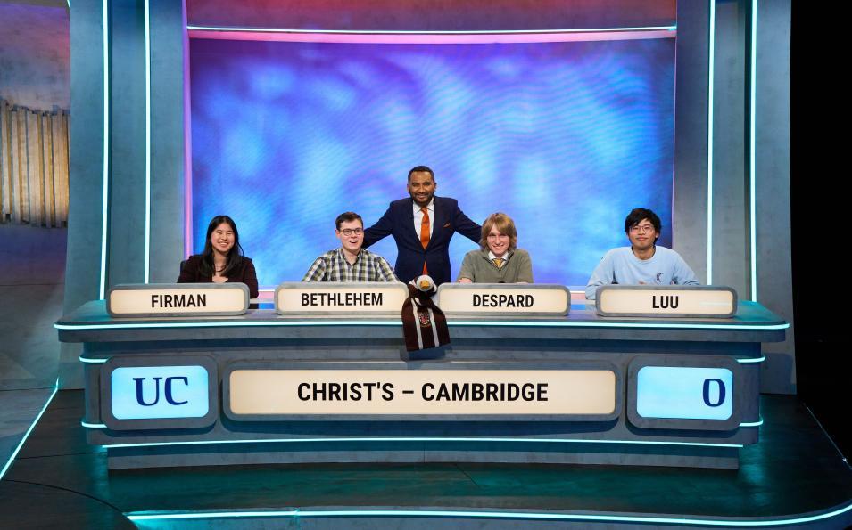 Four people seated in a row in a TV studio, with a fifth person standing behind them