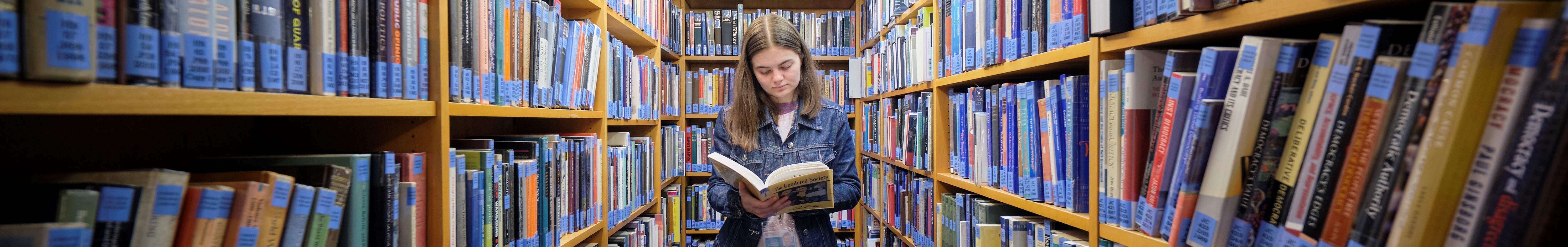 Female, standing with open book, reading between bookshelves.