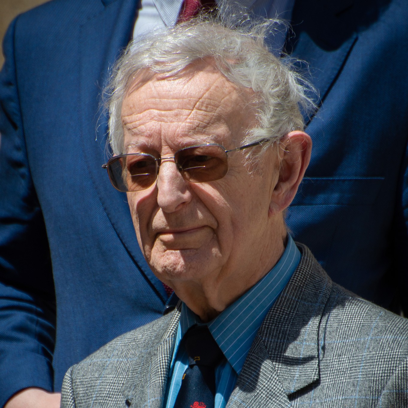 Michael Edwards headshot. He is outside in sunshine and wearing sunglasses, dressed in a grey suit and blue shirt and tie. 