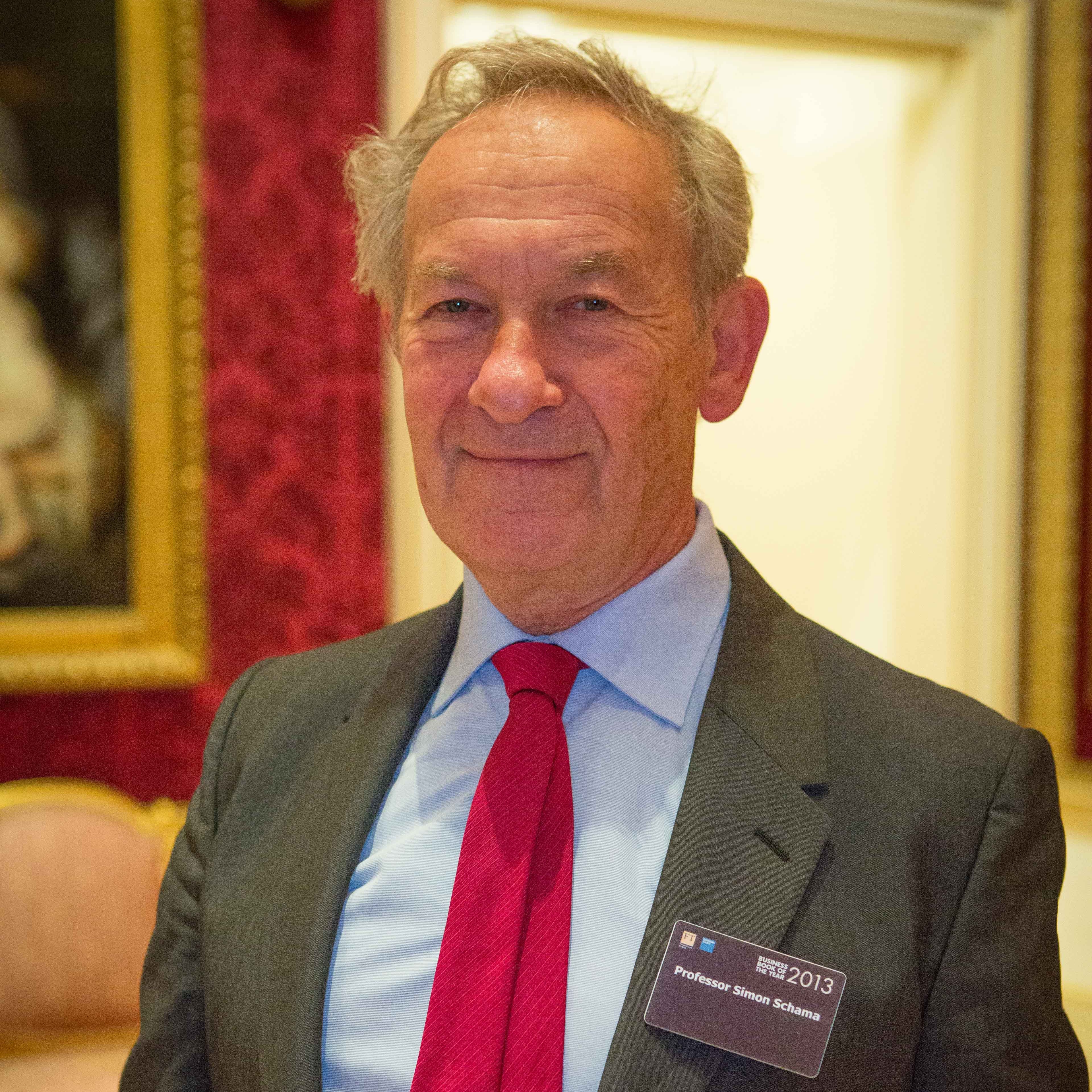 Simon Schama, smiling, wearing grey suit with name badge pinned to lapel, blue shirt and red tie